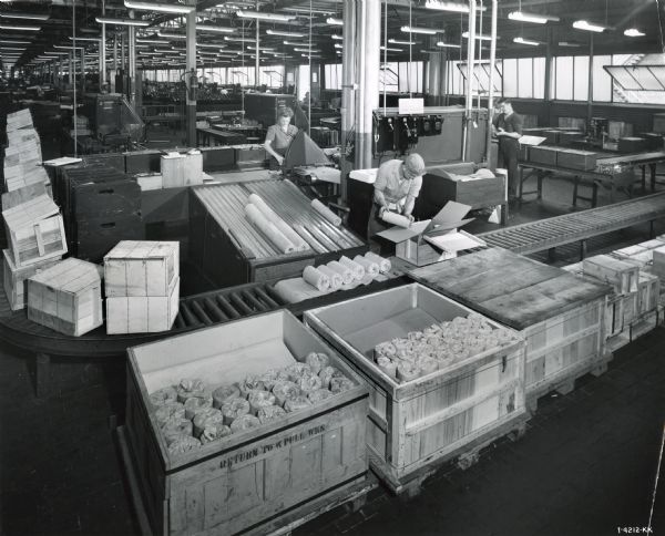 Male and female factory workers at West Pullman Works wrap items in paper and package them in boxes as they roll down a conveyor belt.
