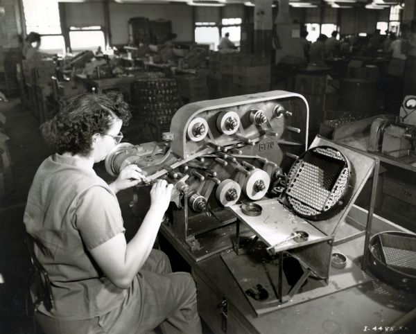 A female factory worker operates a machine consisting of multiple gears while seated at a table at West Pullman Works.