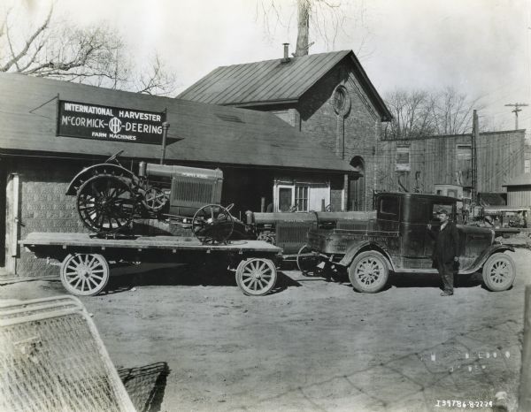 A man stands near a McCormick-Deering (10-20 or 15-30) tractor on a trailer at an International Harvester dealership. The trailer is hitched to an International truck.