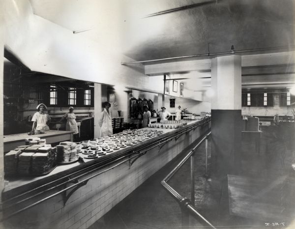 Female cafeteria workers standing behind a counter lined with sandwiches, pies, and coffee cups at International Harvester's Hamilton Works factory.