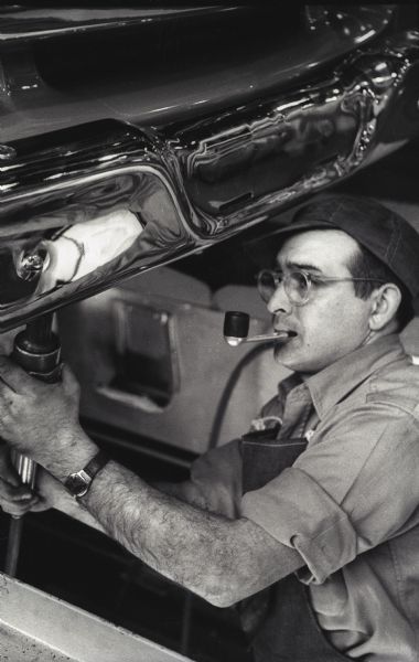 Harlan Riegel installs a chrome bumper on an International A-100 "Golden Jubilee" truck while holding a tool in his mouth at International Harvester's Springfield Works.