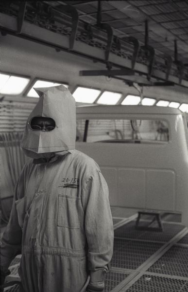 An International A-100 "Golden Jubilee" truck sits on the paint line at Springfield Works. A factory worker wearing a protective suit and hood looks toward the camera.
