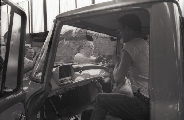 Two factory workers work in the cab of an International A-100 "Golden Jubilee" truck while others wipe the "sweep-around" windshield at International Harvester's Springfield Works. The man sitting at the steering wheel is smoking a pipe.