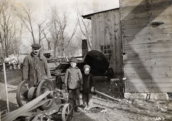 A father standing with his two young sons near a stationary engine. Many farm implements and equipment are lay in the yard.