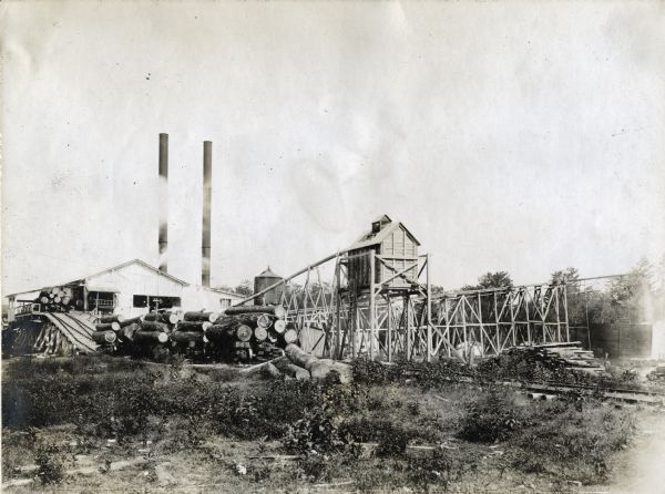 Exterior view of an International Harvester sawmill, with logs stacked on railroad cars on a railroad track.
