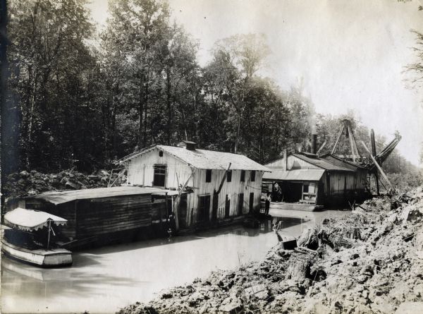 Buildings and boats on water at or near an International Harvester logging camp or sawmill. A man is standing on a crane on the right side of the image, and another man is standing near the water.