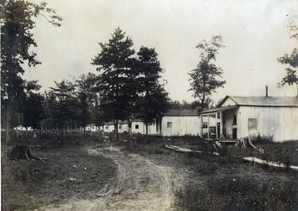 A woman stands in the doorway of the first in a line of houses at an International Harvester logging camp or sawmill.