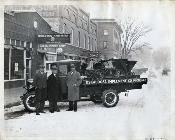 Three men pose with an International "traveling sample floor" truck in front of the Oskaloosa Implement Company building. The original caption reads: "A traveling sample floor.  Middle of winter though it is this salesman takes his samples with him. The Oskaloosa Implement Co., Oskaloosa, Iowa, fitted out this Six-Speed Special with platform body for the special use of the canvasser. It is not used for delivery or any kind of hauling. It is wholly a canvasing (<i>sic</i>) proposition and the truck chassis is to be sold and a new one substituted before there is much if any depreciation in value. The truck is kept continually in the minds of the farmers and the salesman, as well as added interest at each call through the sample machines carried. The men in the picture are, left to right, C.A. Hefley, member of the firm, W.E. Mack, blockman, Scott Hilton, canvaser (<i>sic</i>) for the Implement Co. Mr. Mack is being transferred to foreign sales. We understand that he is to report in Chicago soon and later sail for Singapore."