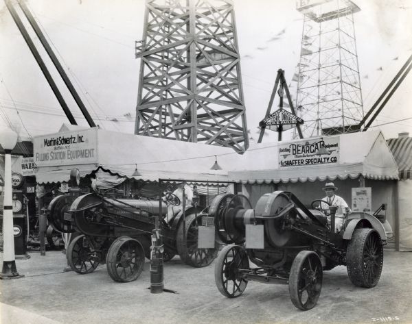 A man sits on a tractor outfitted with a Bear Cat winch or hoisting device near the Schaffer Specialty Company's booth at the International Petroleum Exposition. There are two towers behind the booths.