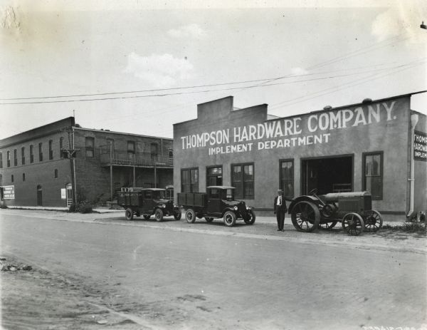 View across street towards a man standing next to two parked trucks and a tractor in front of the Thompson Hardware Company, an International Harvester dealership.