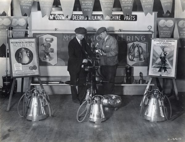 Two men standing amongst McCormick-Deering milking machine parts in a display decorated with signs and overhead banners. The display was most likely part of a state fair, possibly at Milwaukee, Wisconsin.