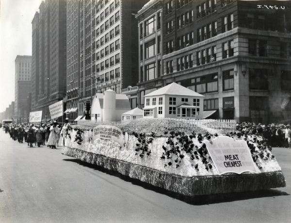A large float decorated with a farm scene and blanketed with flowers moves down a city street (possibly Michigan Avenue in Chicago). The float is part of the Meat Packers Parade, and is followed by marchers holding signs. The banner on the side of the float reads: "Of the total United States wealth, agriculture produces twelve billion dollars annually." The sign on the front reads: "Here is the source of your dinner's main course, and MEAT is the CHEAPEST in years."