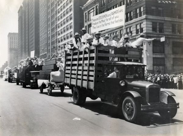 A group of men wave American flags while riding in an International Harvester truck as part of the Meat Packers Parade. The parade is moving down a city street (possibly Michigan Avenue in Chicago). The sign on the truck reads: "These are STOCKMEN and FARMERS who Produce the Meat for all of you; They live on the Farms that feed you."
