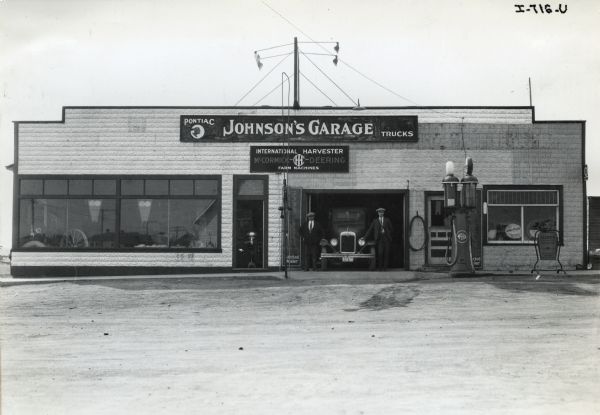 Two men stand on either side of a truck in front of Johnson's Garage, operated by W.E. Johnson. Johnson's Garage was an International Harvester dealership and was located in Oyen, Alberta, Canada. The dealership also includes a "Pontiac" sign and a gas pump.