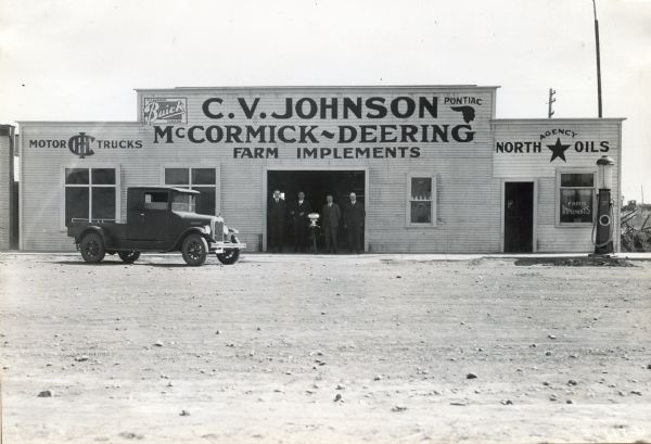 View across lot towards four men posing next to a cream separator in the garage doorway of C.V. Johnson McCormick-Deering Farm Implements, an International Harvester dealership in Hanna, Alberta, Canada. The building also includes signs for "North Oils", "Pontiac", and "Buick" as well as a gas pump.