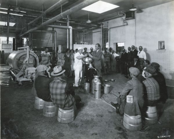 A group of men and women are gathered to watch two men demonstrate a McCormick-Deering cream separator at A&P Creamery. The cream separator was sold by the Pilcher Hardware Company, an International Harvester dealership.