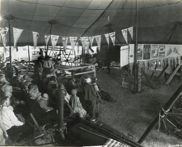 An audience at the Nebraska State Fair sits in the International Harvester tent to watch a motion picture. Banners advertising IH farm equipment and repairs and McCormick-Deering twine, ball-bearing separators, and farm equipment hang from the posts in the tent.