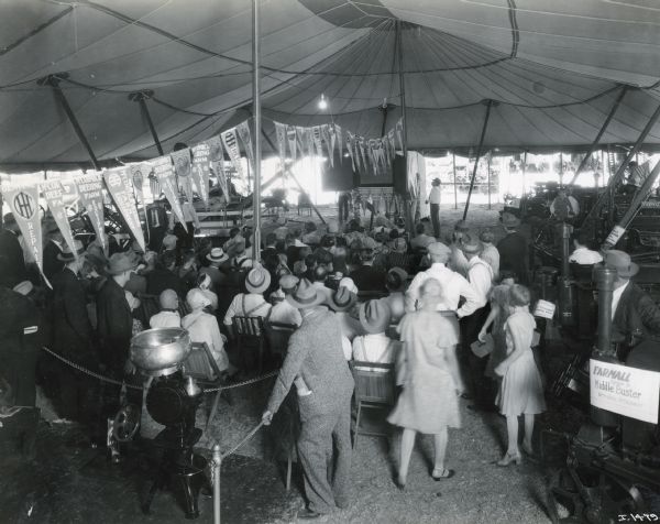 A crowd gathers in the International Harvester tent at the Nebraska State Fair for a film showing. A cream separator is in the left foreground, and next to it stands a Farmall No. 3 Middle Buster with a drill attachment.