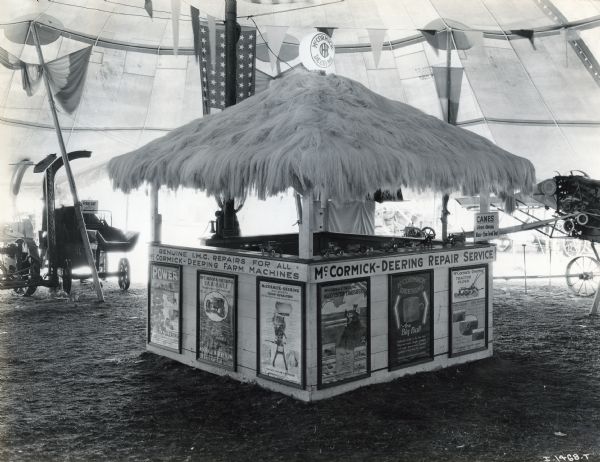 An International Harvester booth with a thatched roof (possibly constructed of binder twine fiber) stands underneath a tent at the Indiana State Fair. There are posters on the sides of the booth, and toy trucks and tractors are displayed above them on the counter. The text on the booth reads: "Genuine I.H.C. Repairs for All McCormick-Deering Farm Machines; McCormick-Deering Repair Service" and "Canes Given Away 10 a.m. and 2 p.m. - Front Tent." The machine in the background at left has a sign reading: "Price Cut for Quick Sale."