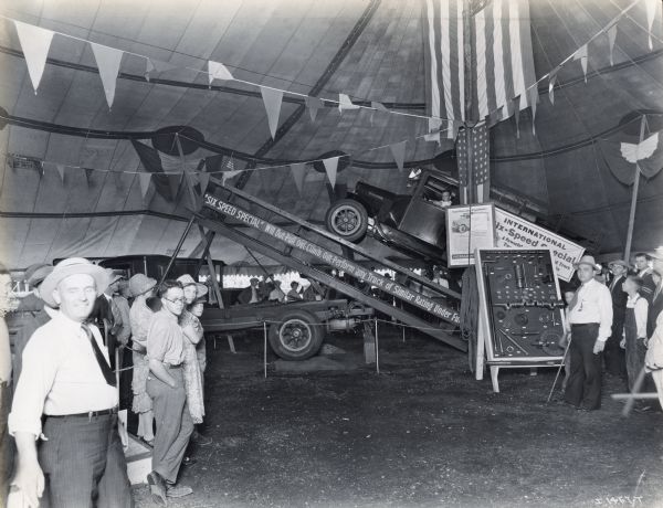 A group of onlookers watches as an International Six-Speed Special truck ascends a ramp in an International Harvester tent at the Indiana State Fair. A case of truck parts is on display to the left. The text on the truck ramp reads: "'Six-Speed Special'; Will Out-Pull, Out-Climb, Out-Perform any Truck of Similar Rating Under Fu..."