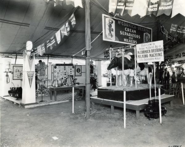 A cow standing on a platform is used to demonstrate a McCormick-Deering milker in the International Harvester tent at the Wisconsin State Fair.  A display board of milker parts is in the background at left. The sign next to the platform reads: "This Cow is Being Milked By a McCormick-Deering Milking Machine - The Dependable Milker."