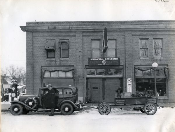 A man, possibly the I.E. Lillegaard, stands next to an automobile pulling a wagon in front of I.E. Lillegaard's International Harvester dealership. A pump for Red Crown Gasoline stands to the left of the building and an advertisement reading: "Red Crown for Power Mileage" is attached next to the garage door.