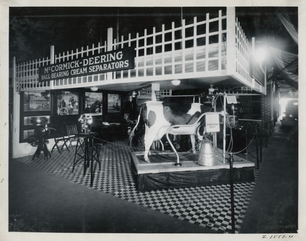 A mechanical(?) cow standing on a platform demonstrates the use of a McCormick-Deering milker at the Dairy Cattle Congress. There are paintings of agricultural life on the wall in the background, and a cream separator on display.