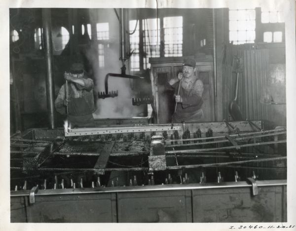 Two factory workers use long-handled tongs to handle a long piece of heated metal at the McCormick Works factory.