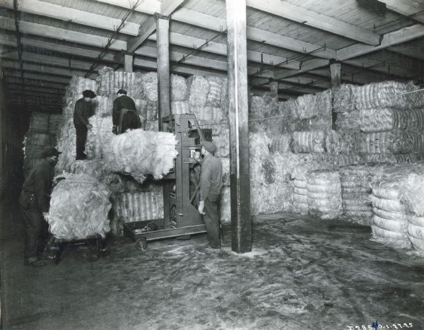 Three men use a lift to stack bales of bound fiber in a storage area, possibly at the McCormick Works twine mill. The company used sisal and hemp fiber to create binder twine.