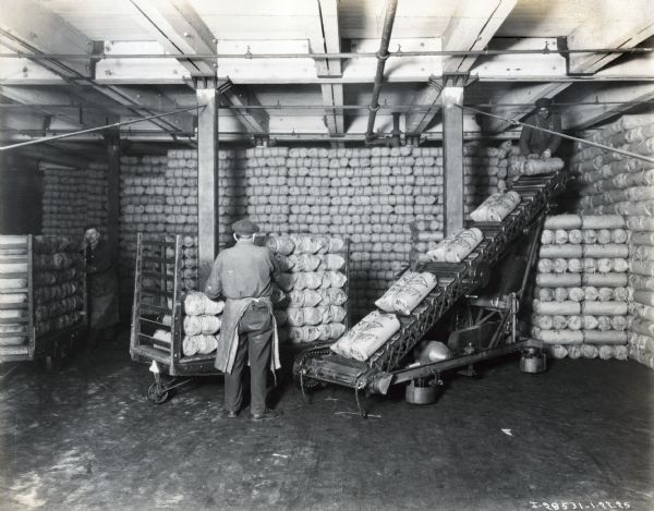 A factory worker loads bags of twine onto a conveyor belt, while another waits at the top of the pile to stack the twine in a storage space, possibly at the McCormick Works twine mill.