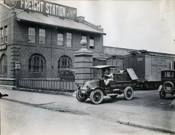 An International truck labeled "Kingston's Baggage Line" and piled with luggage trunks pulls out of the parking lot of a Northwestern Line Freight Station. The truck is likely a model H, or 21.