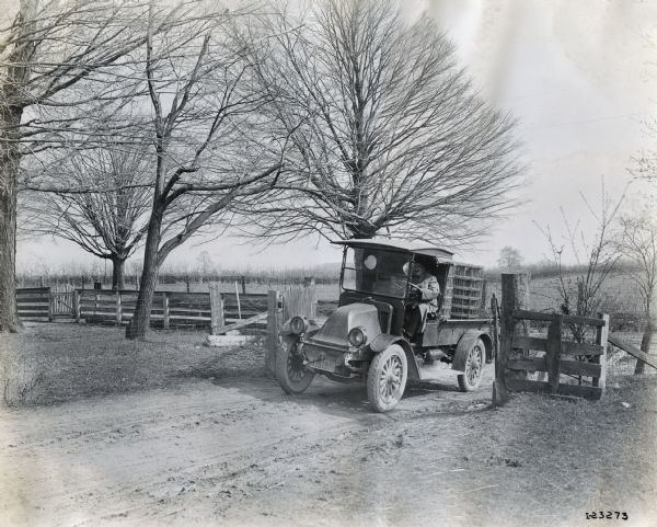 A man drives an International truck down a dirt road near a fence. The truck is loaded with what appear to be coils of wire fencing. The truck is likely a model H, or 21.