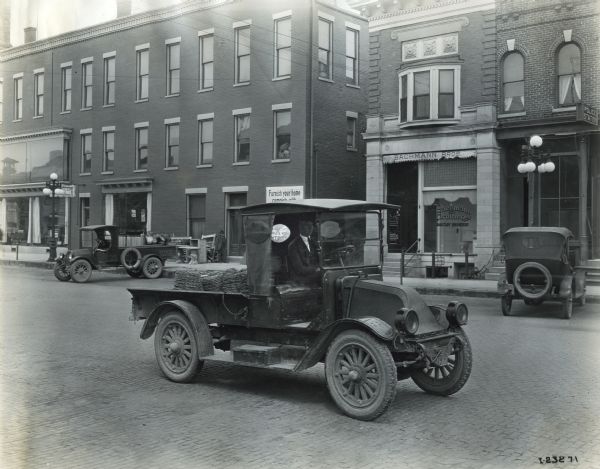 An International truck carries goods down a street past the storefronts of Kesslers' Furniture Store, Bachmann Brothers Sanitary Engineers Shop, and a police station. The truck is likely a model H, or 21.