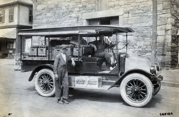 Two men pose with an International truck loaded with Moxie soft drink bottles parked in front of a stone building. The text on the bottle containers reads: "Drink Moxie" and "Bachelor's Bottling Works." The truck is likely a model H, or 21.