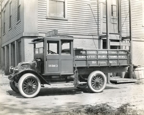 International truck used by Milford Furniture Company parked in front of a building. The text on the truck reads: "Milford Furniture Co. Complete Home Furnishers; Milford, Mass.," "Glenwood Ranges," and "128 Main St." The truck may be a Model H or 21.