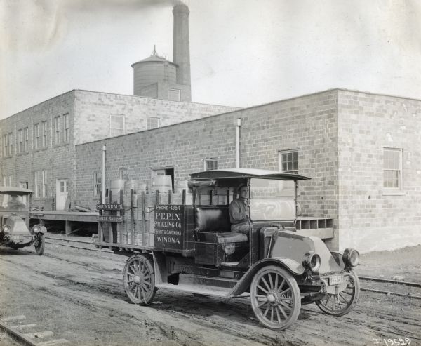 A man driving an International truck for Pepin Pickling Company. The truck is loaded with boxes of sliced dill and mixed sweet pickles, and traveling down a dirt road next to a brick building with a loading dock and railroad tracks. The truck may be an International Model H or 21.