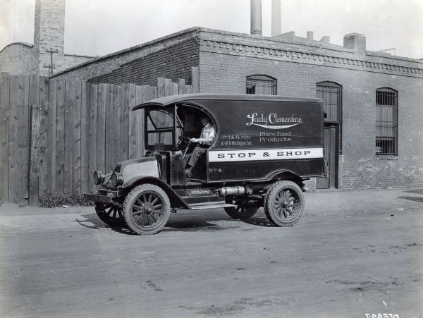Man driving an International truck advertising "Lady Clementine Pure Food Products" is parked on a dirt road in front of a fence near a brick building. The truck may be a mode "H" or 21.