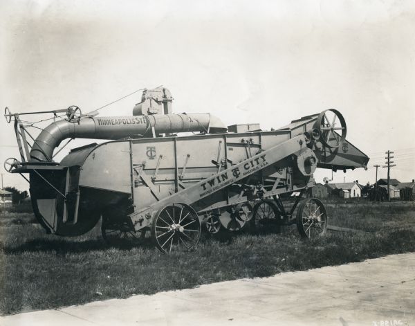 Twin City threshing machine in grassy area with houses in the background. The machine bears the text: "Twin City; Minneapolis Steel & Machinery Co., Minneapolis, U.S.A."