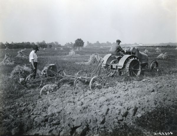 Two farmers use an International 8-16 tractor with attached machinery to plow a farm field. Harvested bundles of the crop are in the distance.