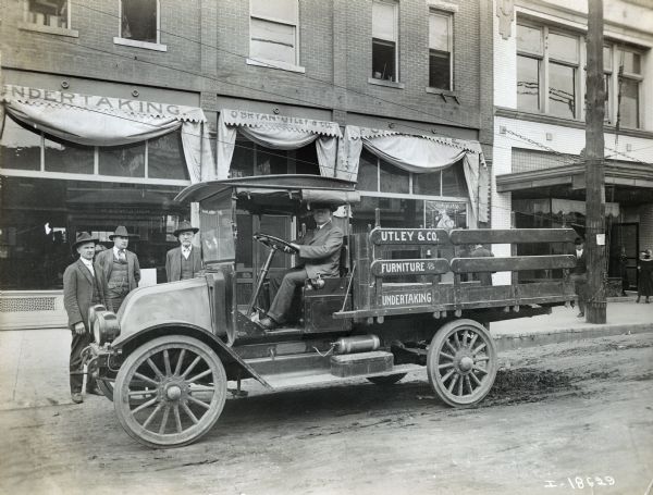 Men standing near an International truck used by the Utley & Co., a furniture and undertaking company. The Utley & Co. storefront is in the background. The truck may be a Model H or 21.
