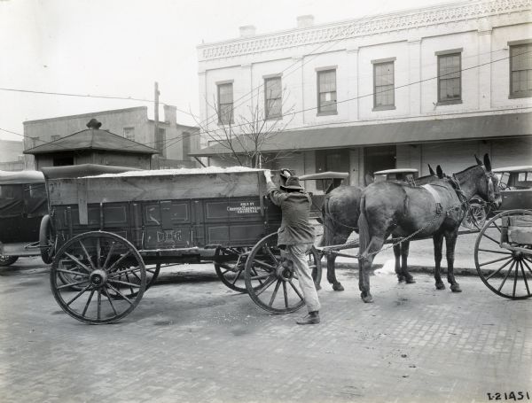 A man climbs onto a Weber wagon led by two mules parked in an area behind several buildings. Autombobiles are in the background. The text on the wagon reads, "Weber; King of All" and "Sold by Griffin Hardware Co., Griffin, Ga." The Weber Wagon Company was started by Henry Weber in 1845 and was acquired by the International Harvester Company in 1904.