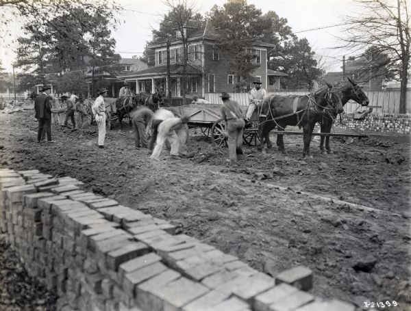 Men use mule-led wagons while working on a residential road.  Bricks are piled high on either side of the street.