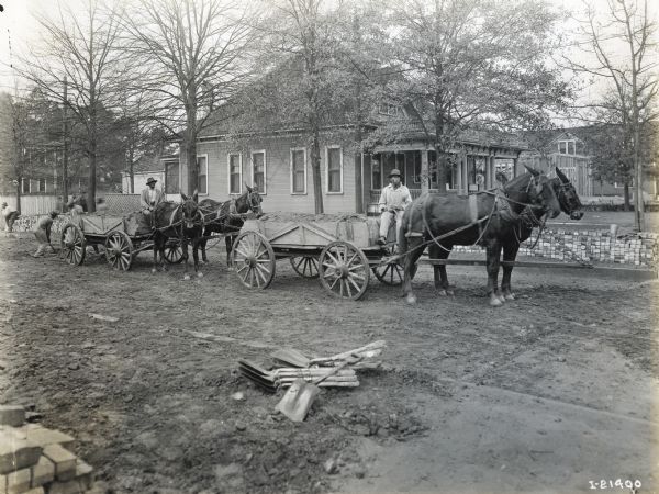 Men using mule-drawn wagons as they prepare to pave a street with bricks. A pile of shovels is lying in the dirt in the foreground.