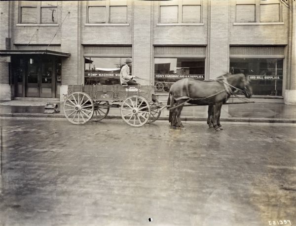 A man driving a Weber wagon with two mules sitting in front of a building in front of the Komp Machine Works.
