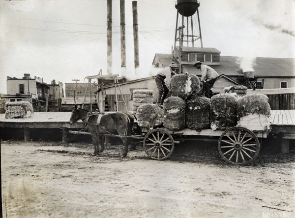 Two men place a bale of cotton on top of a pile on the back of a mule-drawn wagon. They are near what appears to be a weighing station on a wooden platform. There are smokestacks and a water tower in the background.