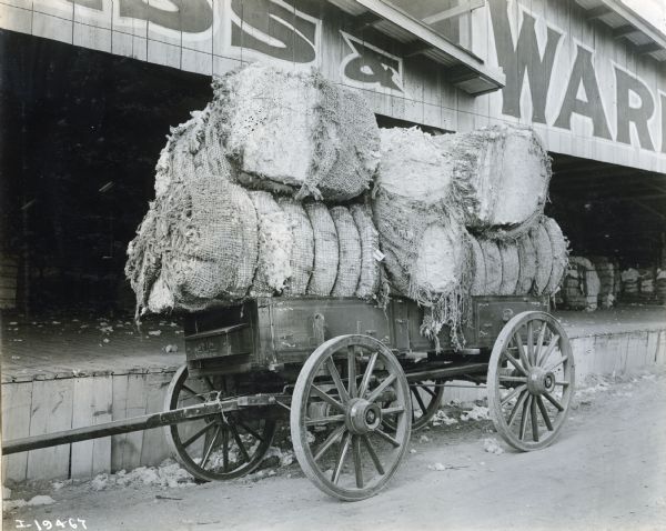 A wagon loaded with bales of cotton sits in front of a loading dock.  The wagon was possibly made by the Weber Wagon Company.