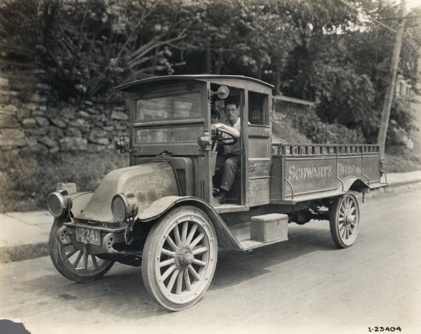 International model "F" or "31" truck operated by "Shwartz Bros. Inc." of "Norwich Conn." Two men, a driver and a passenger, are sitting in the truck. Additional advertising on the truck reads: "The Big Store With The Little Prices."