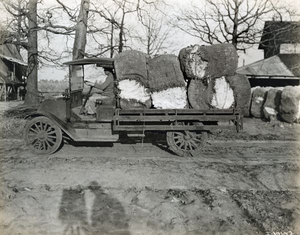 International model "F" or "31" truck. The truck contains bales of cotton, and a man sits in the driver's seat. The truck is photographed on a dirt road with wooden work(?) buildings in the background.  Additional cotton bales can be seen in the background. The shadow of the photographer is in the foreground.