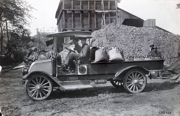 International model "F" or "31" truck carrying bags of what appears to be gravel. Two men, a driver and passenger, are sitting in the truck. In the  background is a horse, a wooden structure, and two men (one with a shovel), in front of pile of gravel(?).