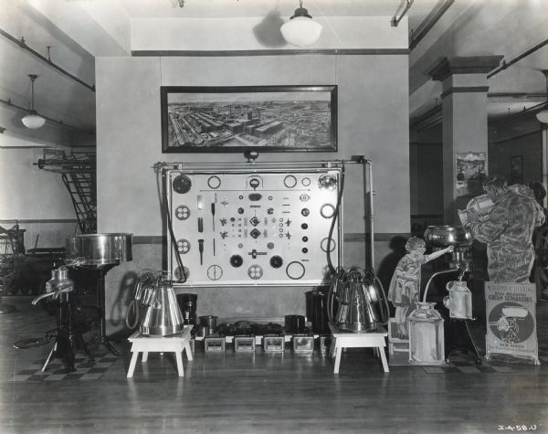 Ball bearing cream separators and milkers are arranged in a display in a hallway at International Harvester's Milwaukee Works factory. A showcase of individual machine parts hangs on the wall underneath a bird's-eye view painting of the McCormick Works factory.